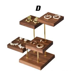 May include: A wooden jewelry stand with four tiers. The stand is made of dark wood and has gold metal posts. The top tier has two heart-shaped earrings, the second tier has two geometric earrings, the third tier has two hoop earrings and a ring, and the bottom tier has a ring and a bracelet.