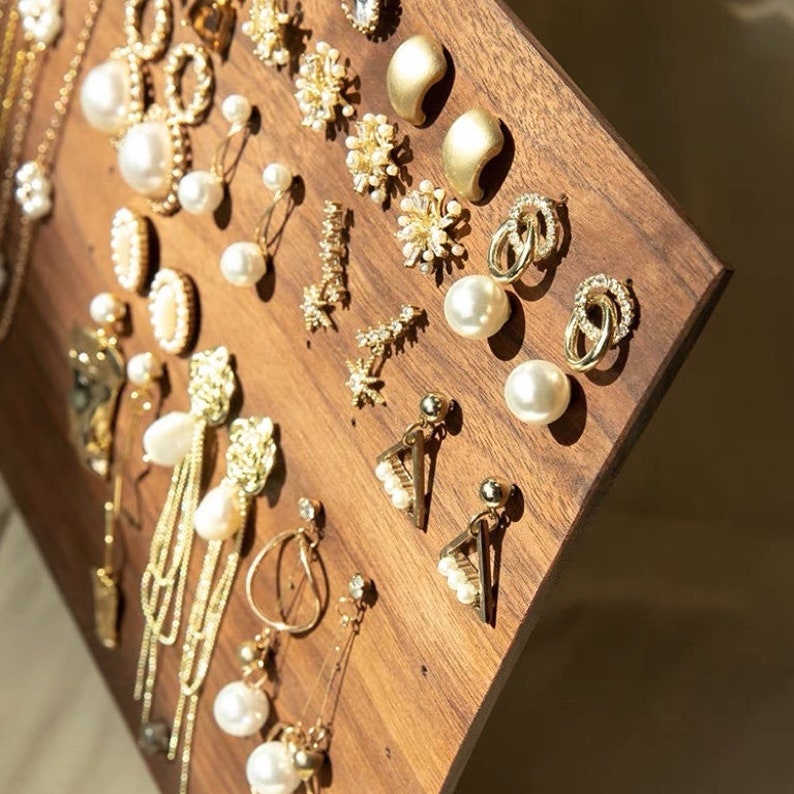Puede incluir: Un tablero de exhibici&oacute;n de aretes de madera con una variedad de aretes de oro y perlas. Los aretes est&aacute;n dispuestos en filas y columnas, y algunos tienen dijes colgantes.