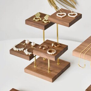 May include: A tiered wooden jewelry stand with gold metal rods. The stand has four square platforms, each displaying different jewelry pieces, including earrings, rings, and necklaces.