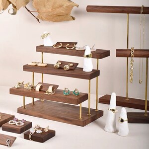 May include: A tiered wooden jewelry display stand with brass rods and white ceramic ring holders. The stand has three levels for displaying earrings, rings, and necklaces. The stand is made of dark walnut wood and has a modern design.