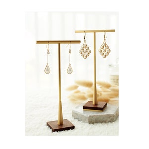 Op de afbeelding: Twee gouden metalen oorkringstandaards met houten bases. De standaards hebben een T-vormige balk om oorkringen te houden. Eén standaard heeft twee paar parel-druppel oorkringen, de andere heeft een paar parel-cluster oorkringen.