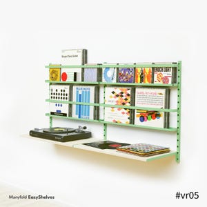 Puede incluir: Una estantería de pared verde menta que muestra discos de vinilo y CD. Un tocadiscos negro se encuentra en un estante inferior. Las estanterías contienen varias portadas de álbumes con diseños coloridos. El texto "Manyfold EasyShelves" y "#vr05" son visibles.