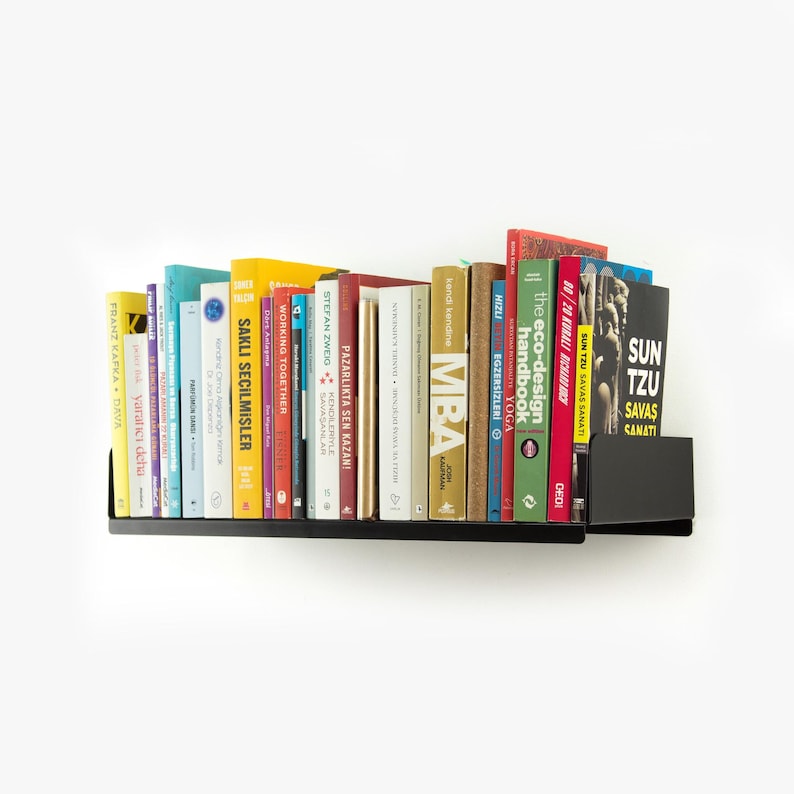 May include: A black metal floating shelf with a variety of books, including "The Eco-Design Handbook", "MBA", "Sun Tzu Savas Sanati", and "Pazarlikta Sen Kazan!" The shelf is mounted on a white wall.