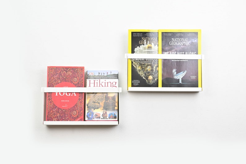 May include: Two white metal magazine holders mounted on a white wall. The top holder has two yellow National Geographic magazines. The bottom holder has three books: a red book with the word "YOGA" on the cover, a book with the word "Hiking" on the cover, and a book with a black and white photo of a cat on the cover.