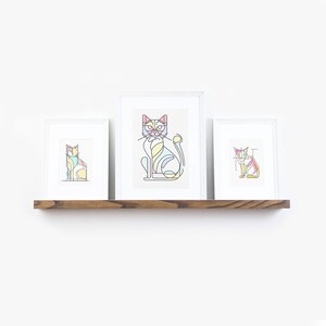 May include: Three framed cat art prints with a geometric design in pastel colors. The prints are displayed on a dark wooden shelf against a white wall. The cat illustrations feature a variety of colors, including pink, yellow, and blue.