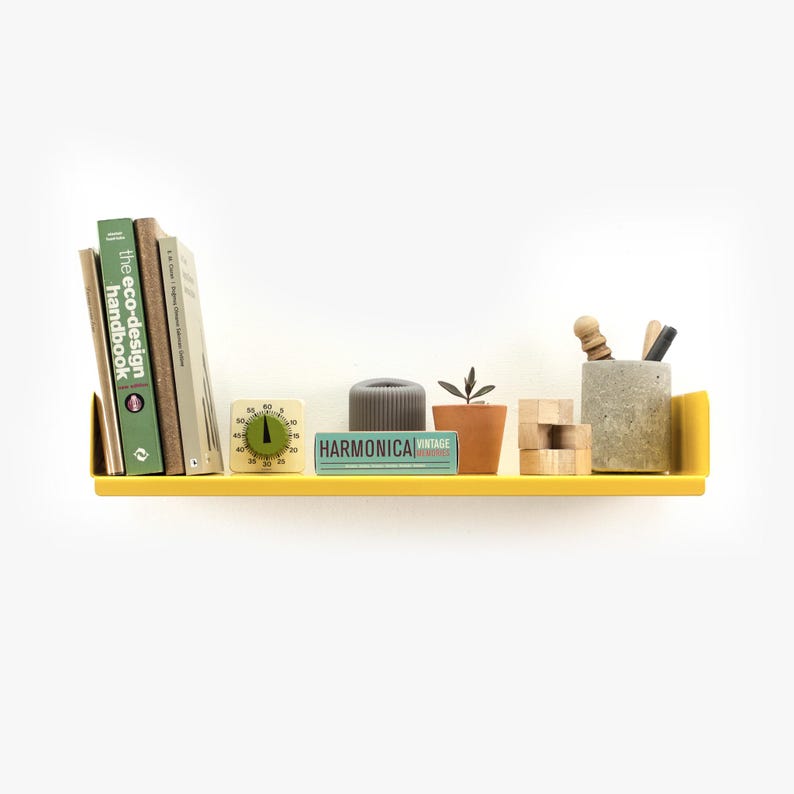 May include: A yellow metal shelf with a variety of items on it, including books, a small plant, a timer, a candle, and a pencil holder. The shelf is mounted on a white wall.