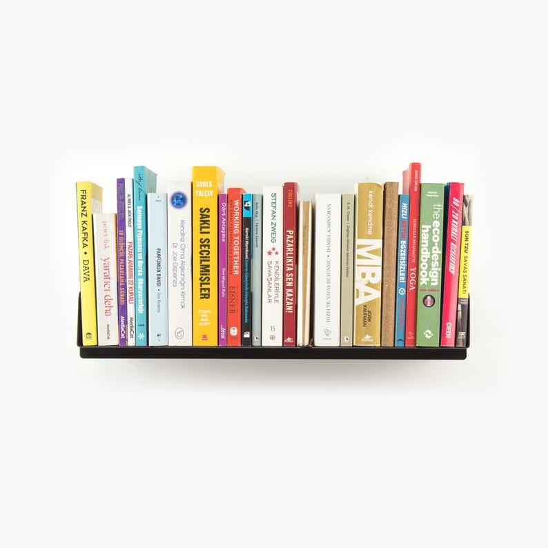 May include: A black metal shelf with a variety of colorful books. The books are arranged in a row and include titles such as "Working Together", "MBA", "The Eco-Design Handbook", and "Sun Tzu Savas Sanati".