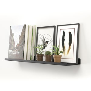 May include: A black metal floating shelf mounted on a white wall, displaying books, framed art, and potted plants. The art includes a geometric bird design and feather illustrations. The book on the left has the word "Wroclaw" on the cover.
