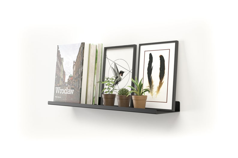 May include: A black metal floating shelf mounted on a white wall, displaying books, framed art, and potted plants. The art includes a geometric bird design and feather illustrations. The book on the left has the word "Wroclaw" on the cover.