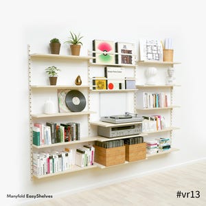 Può includere: Una mensola a muro in legno chiaro con libri, dischi in vinile, piante e un giradischi. I ripiani sono fissati a una parete bianca. Il testo "Manyfold EasyShelves" e "#vr13" sono visibili. Una soluzione di archiviazione elegante.