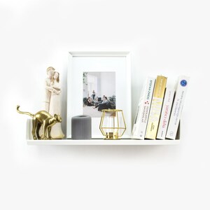 May include: A white shelf with a gold metal cat figurine, a gray cylindrical candle, a gold metal cage candle holder, a framed photo, and a stack of books. The books have titles in Polish and German.
