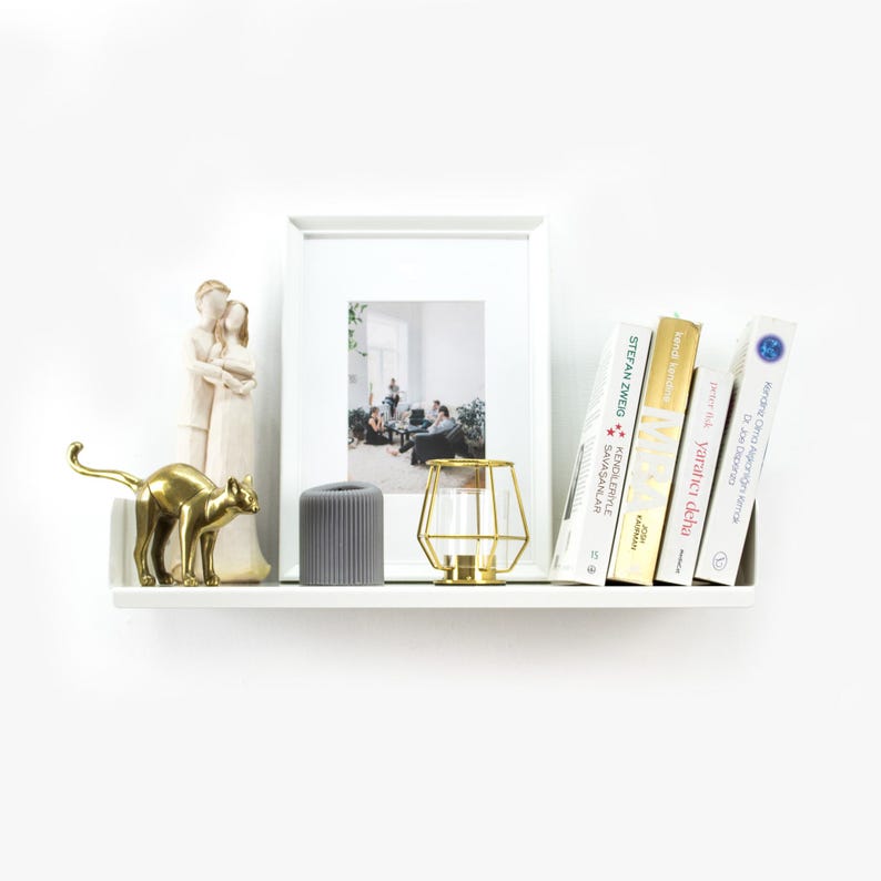 May include: A white shelf with a gold metal cat figurine, a gray cylindrical candle, a gold metal cage candle holder, a framed photo, and a stack of books. The books have titles in Polish and German.