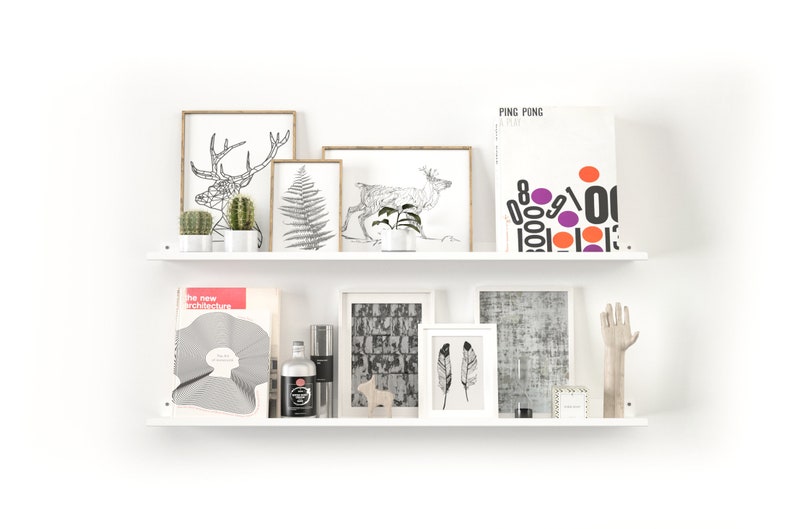 May include: Two white shelves displaying framed art, plants, and decorative objects. The top shelf features botanical prints and a graphic poster. The bottom shelf holds a book, a bottle, and a wooden hand sculpture. The overall aesthetic is minimalist and modern.