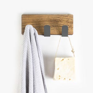 May include: A wooden wall-mounted hook rack with three black metal hooks. A gray and white striped towel hangs on one hook and a bar of soap hangs from another hook.