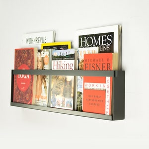 May include: A black metal magazine rack mounted on a white wall with various magazines and books on the shelves. The magazines include titles like "WOHNREVUE", "edare", "Hiking", "Homes & Gardens", "Working Together", and "TOGA".