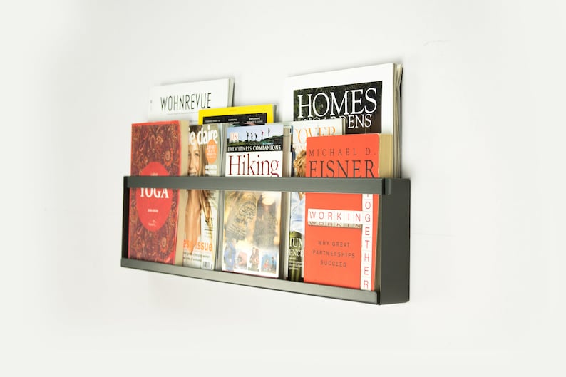 May include: A black metal magazine rack mounted on a white wall with various magazines and books on the shelves. The magazines include titles like "WOHNREVUE", "edare", "Hiking", "Homes & Gardens", "Working Together", and "TOGA".