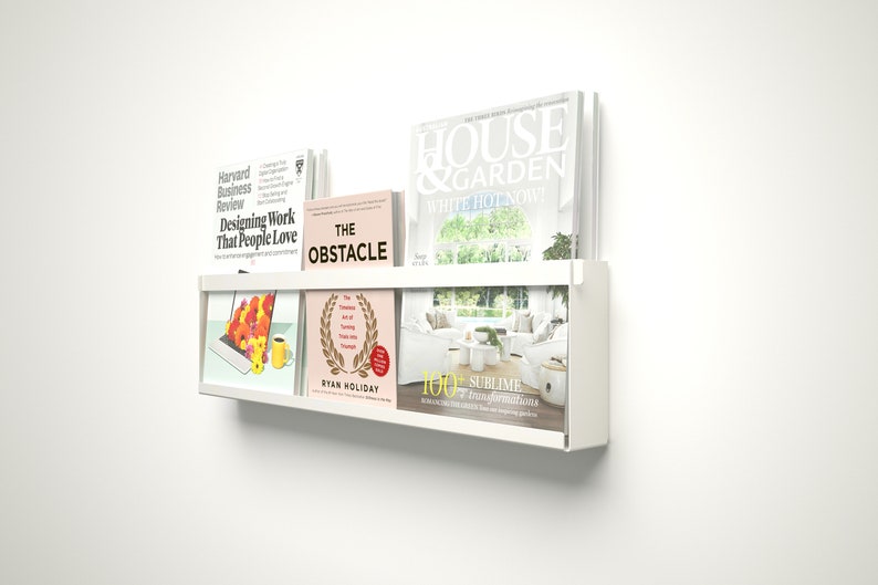 May include: White wall mounted shelf with two tiers holding various books and magazines. The top tier has a magazine titled "House & Garden" with the words "White Hot Now" on the cover. The bottom tier has a book titled "The Obstacle" by Ryan Holiday and a magazine with the words "100+ Sublime" on the cover.