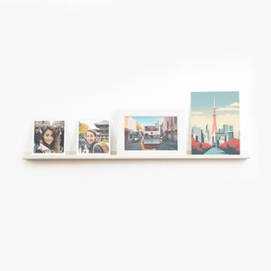 May include: Three white picture frames with photos of a woman in Japan, a woman taking a photo of a street scene, and a cityscape with a tower. The frames are on a white shelf against a white wall.