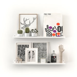 May include: Two white floating shelves with decorative items. The top shelf holds framed art, cacti, and a book titled "PING PONG A PLAY". The bottom shelf displays framed art, a bottle, a hand sculpture, and other decorative objects.