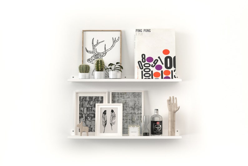 May include: Two white floating shelves with decorative items. The top shelf holds framed art, cacti, and a book titled "PING PONG A PLAY". The bottom shelf displays framed art, a bottle, a hand sculpture, and other decorative objects.