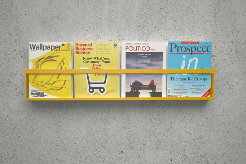 May include: A yellow metal magazine rack with four magazines on it. The magazines are "Wallpaper*" by Zaha Hadid, "Harvard Business Review", "Politico", and "Prospect".