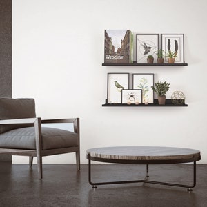 May include: A modern living room scene featuring a gray armchair, a round coffee table, and black shelves with framed art and plants. The coffee table has a dark metal frame and a wood-grain top. The shelves display art prints and potted plants.