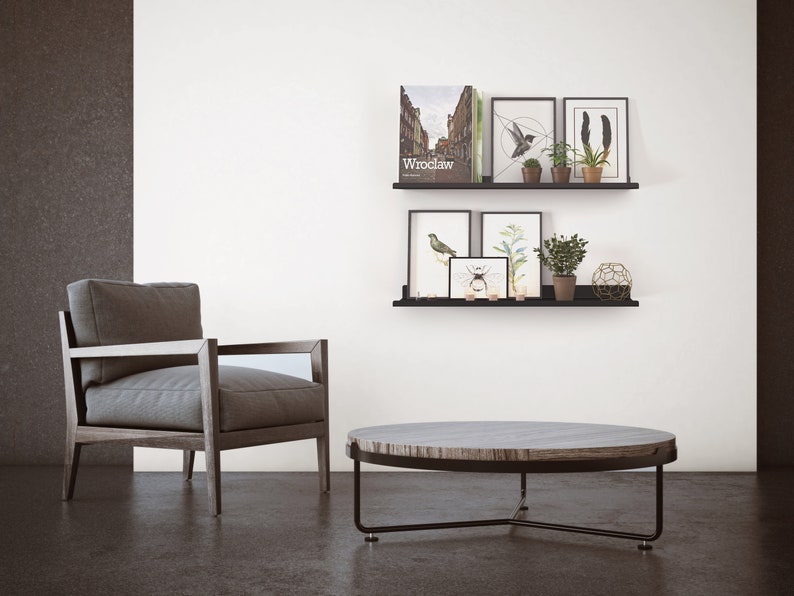 May include: A modern living room scene featuring a gray armchair, a round coffee table, and black shelves with framed art and plants. The coffee table has a dark metal frame and a wood-grain top. The shelves display art prints and potted plants.