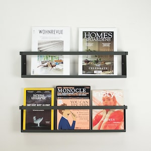 May include: Two black metal magazine racks mounted on a white wall. The top rack holds two magazines, one titled "WOHNREVUE" and the other titled "HOMES & GARDENS". The bottom rack holds four magazines, one titled "NATIONAL GEOGRAPHIC", one titled "MONOCLE", one titled "InStyle", and one with a yellow cover.