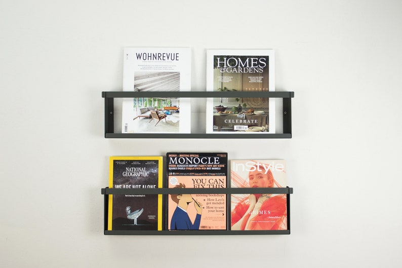 May include: Two black metal magazine racks mounted on a white wall. The top rack holds two magazines, one titled "WOHNREVUE" and the other titled "HOMES & GARDENS". The bottom rack holds four magazines, one titled "NATIONAL GEOGRAPHIC", one titled "MONOCLE", one titled "InStyle", and one with a yellow cover.