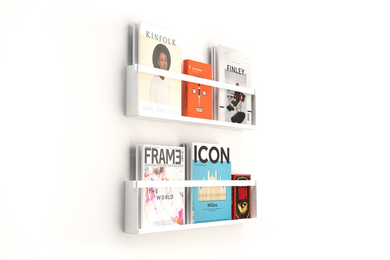 May include: Two white wall-mounted magazine racks with a variety of magazines and books. The top rack has a magazine titled "Kinfolk", a red book with a face on the cover, and a magazine titled "Finley". The bottom rack has a magazine titled "FRAME", a magazine titled "ICON", and a red book with a black and white image on the cover.