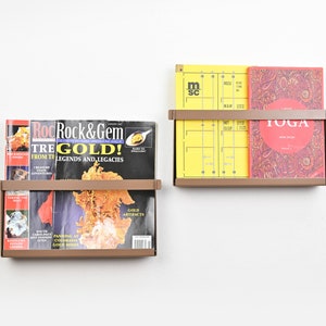 May include: Two brown metal magazine holders mounted on a white wall. The top holder has a yellow magazine with the text "m sc" and a red book with the text "YOGA". The bottom holder has two magazines with the text "Rock & Gem" and "TREGOLD!"