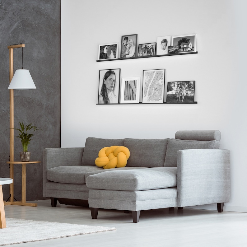 May include: A gray sectional sofa with a yellow knotted pillow. The sofa is in a living room with a white wall and a shelf with black and white framed photos.