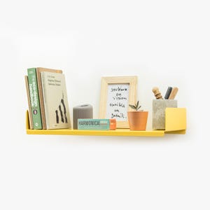 May include: A yellow metal shelf with a rounded edge, holding a stack of books, a small plant in a terracotta pot, a gray concrete pencil holder with pencils, a small gray decorative object, and a small blue and orange box with the word "HARMONICA" printed on it. A framed white board with the text "Stubborn on vision flexible on details." is also on the shelf.