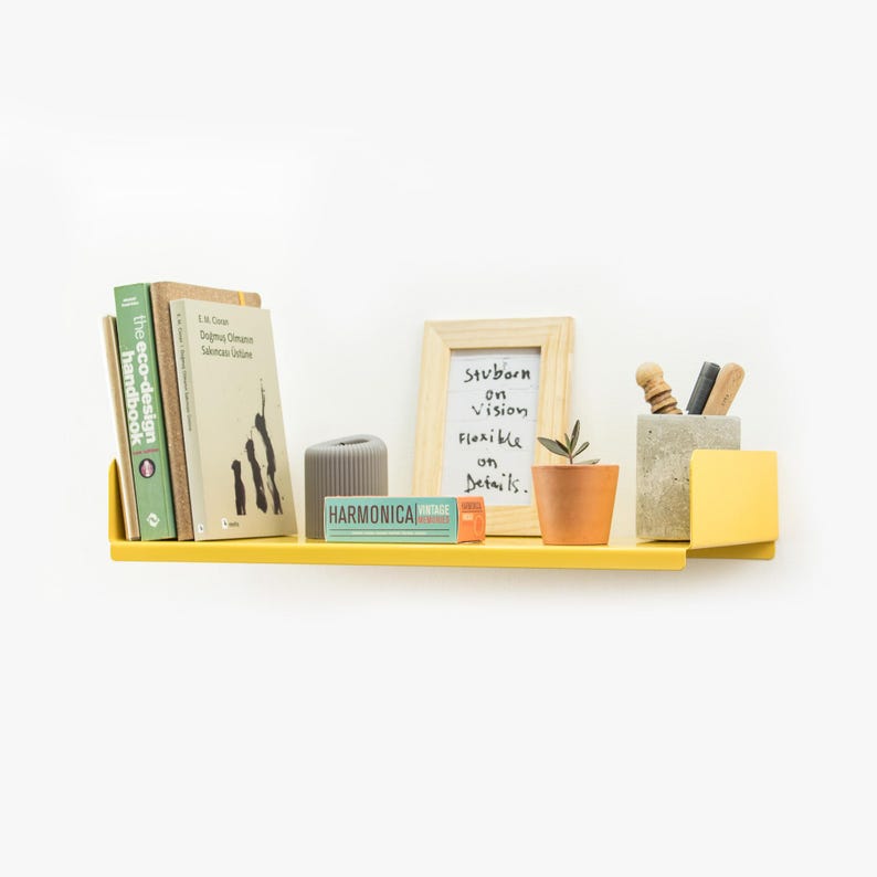 May include: A yellow metal shelf with a rounded edge, holding a stack of books, a small plant in a terracotta pot, a gray concrete pencil holder with pencils, a small gray decorative object, and a small blue and orange box with the word "HARMONICA" printed on it. A framed white board with the text "Stubborn on vision flexible on details." is also on the shelf.