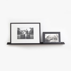 May include: Two black picture frames with black and white photos are displayed on a black floating shelf against a white wall.