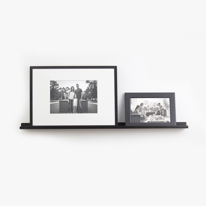 May include: Two black picture frames with black and white photos are displayed on a black floating shelf against a white wall.