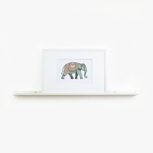May include: A white framed print of a teal elephant with colorful patterned decorations on its back. The elephant is walking to the right. The print is on a white shelf.