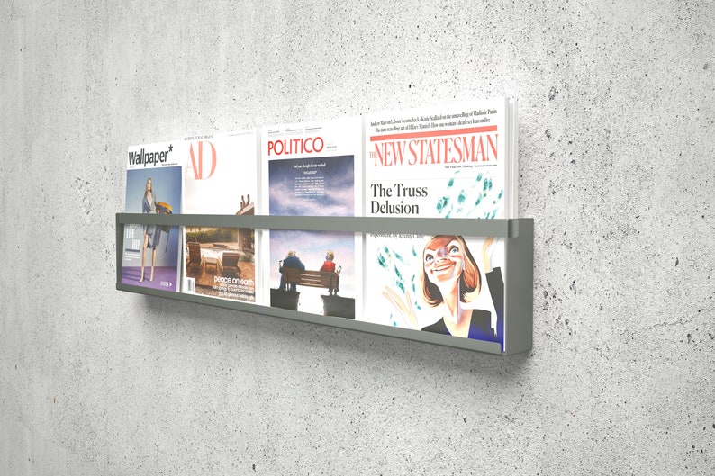 May include: A gray metal magazine rack with four magazines on a concrete wall. The magazines are "Wallpaper*," "AD," "Politico," and "The New Statesman." The "The Truss Delusion" headline is visible on the "The New Statesman" magazine.