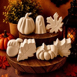 May include: A collection of seven unpainted white ceramic figures featuring seasonal shapes including pumpkins, a ghost, an acorn, and maple leaves. These matte white decorative items are arranged on two stacked wooden platforms for display.