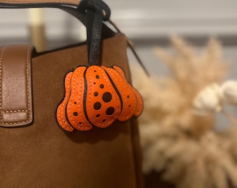 May include: An orange pumpkin-shaped leather bag charm with black dot details, attached to a brown suede handbag. The charm has a black leather strap and is a seasonal accessory.