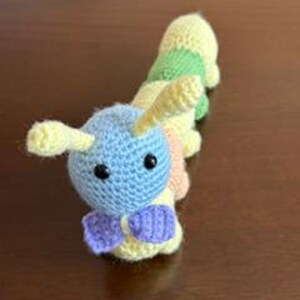 May include: A crocheted caterpillar toy featuring a light blue head with two yellow antennae and black bead eyes. The body is composed of segmented sections in alternating colors of yellow, green, and peach, with a small purple bow attached at the neck.