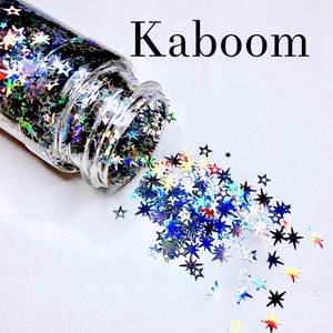 May include: A clear glass jar filled with iridescent star-shaped glitter, spilling out onto a white surface. The glitter is a mix of blue, silver, red, and gold colors. The word "Kaboom" is written in black text above the jar.