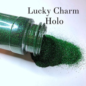 May include: A clear jar of green glitter with a holographic finish. The jar is tipped over and the glitter is spilling out onto a white surface. The text "Lucky Charm Holo" is written above the jar.