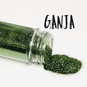 May include: A small clear jar with a green glitter inside. The jar is tipped over and the glitter is spilling out onto a white surface. The word "GANJA" is written in black above the jar.