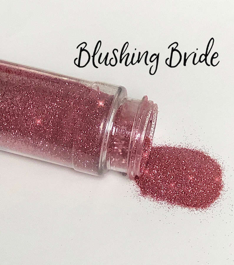May include: A clear plastic jar with a pink glitter inside. The jar is tipped over and the glitter is spilling out. The text "Blushing Bride" is written above the jar.