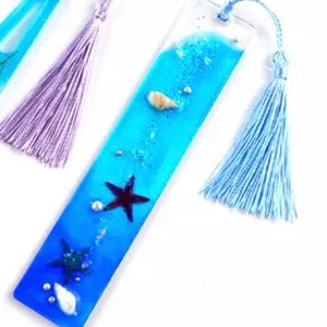 May include: A blue glass bookmark with a seashell, starfish, and glitter embedded inside. The bookmark has a light blue tassel.