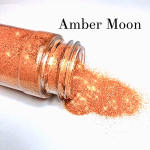May include: A clear plastic jar with a gold glitter inside. The jar is tipped over and the glitter is spilling out onto a white surface. The text "Amber Moon" is written above the jar.