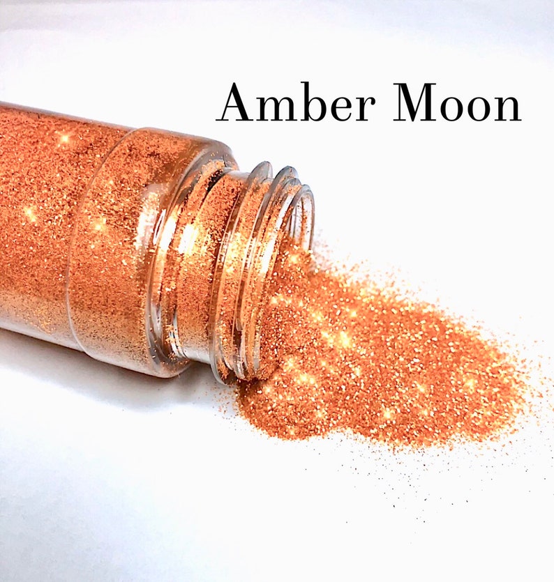 May include: A clear plastic jar with a gold glitter inside. The jar is tipped over and the glitter is spilling out onto a white surface. The text "Amber Moon" is written above the jar.