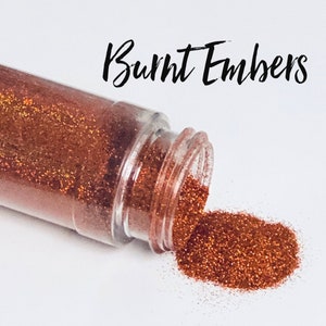 May include: A small jar of loose copper glitter with a gold shimmer. The glitter is spilling out of the jar onto a white surface. The text "Burnt Embers" is written above the jar.