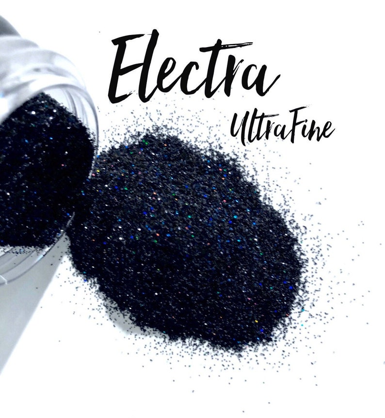 May include: Black glitter spilling from a clear container onto a white surface. The glitter is labeled "Electra Ultra Fine" in black script. The glitter has a dark, almost navy blue appearance with hints of iridescent color.